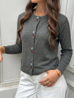 A woman wearing a gray knit cardigan with colorful buttons, paired with white pants, standing against a white paneled door.
