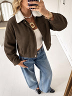 A woman takes a mirror selfie wearing a brown corduroy jacket with a white sherpa collar, styled with a white crop top, wide-leg blue jeans, and black polka-dot shoes.