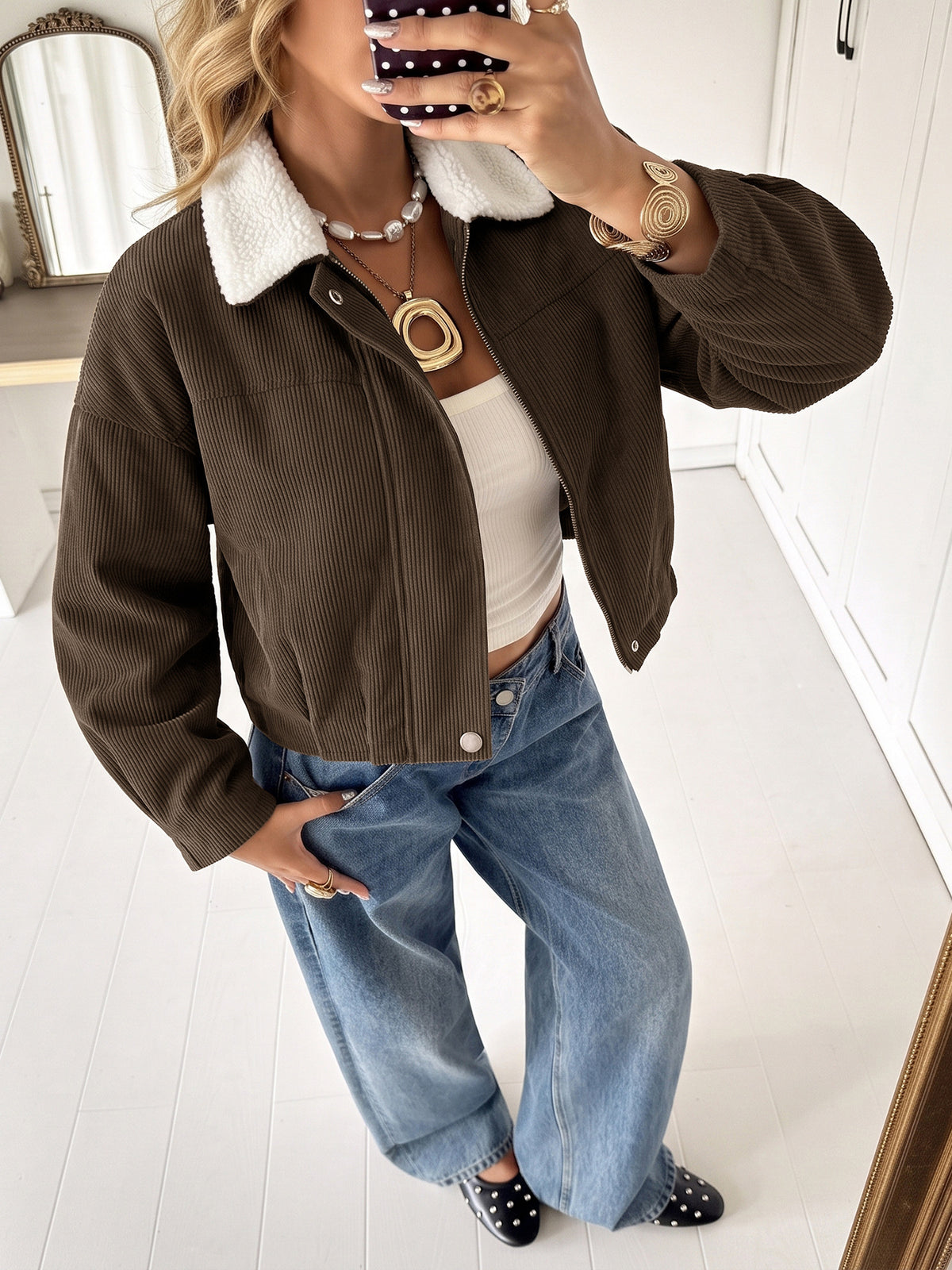 A woman takes a mirror selfie wearing a brown corduroy jacket with a white sherpa collar, styled with a white crop top, wide-leg blue jeans, and black polka-dot shoes.