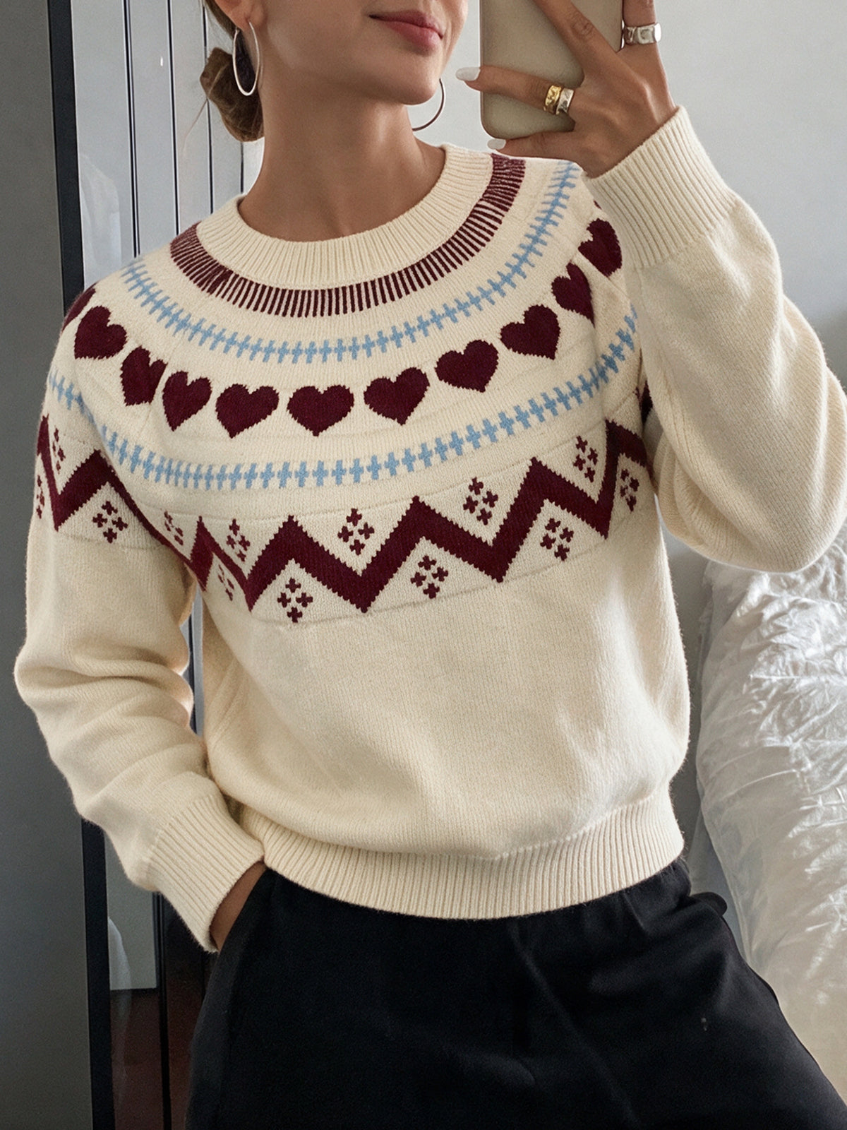 A woman wearing a cream-colored knit sweater with a Fair Isle pattern featuring dark red hearts and zigzags, light blue stripes, and dark brown ribbed trim, paired with black pants.