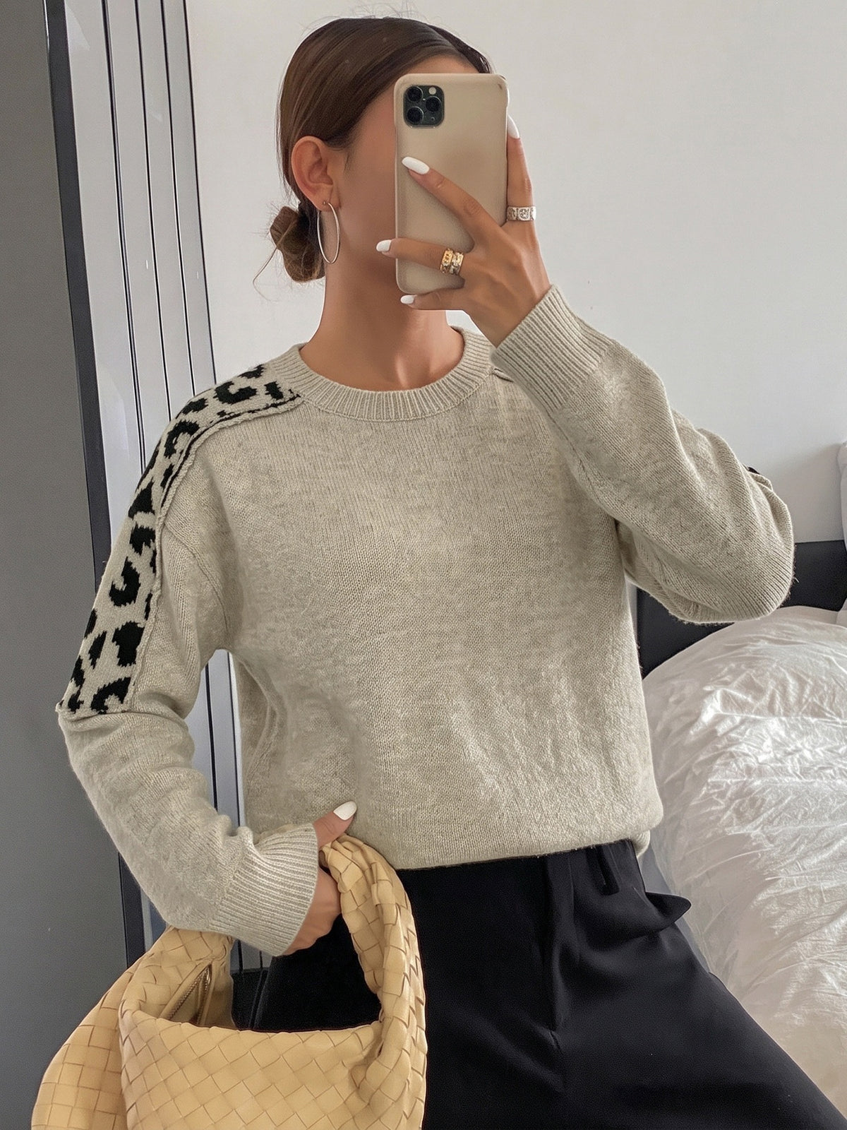 A woman takes a mirror selfie wearing a light beige crewneck sweater with black leopard print knit shoulder panels, paired with black pants and holding a woven beige handbag.