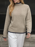A woman wearing a beige knit mock-neck sweater with dark navy blue trim at the collar, cuffs, and hem, paired with white trousers.