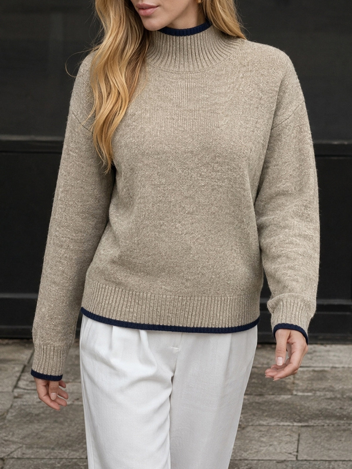 A woman wearing a beige knit mock-neck sweater with dark navy blue trim at the collar, cuffs, and hem, paired with white trousers.