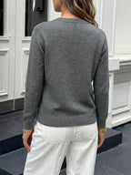 A woman viewed from the back wearing a heather gray long-sleeve knit sweater and white pants, standing outdoors in front of a white paneled building.