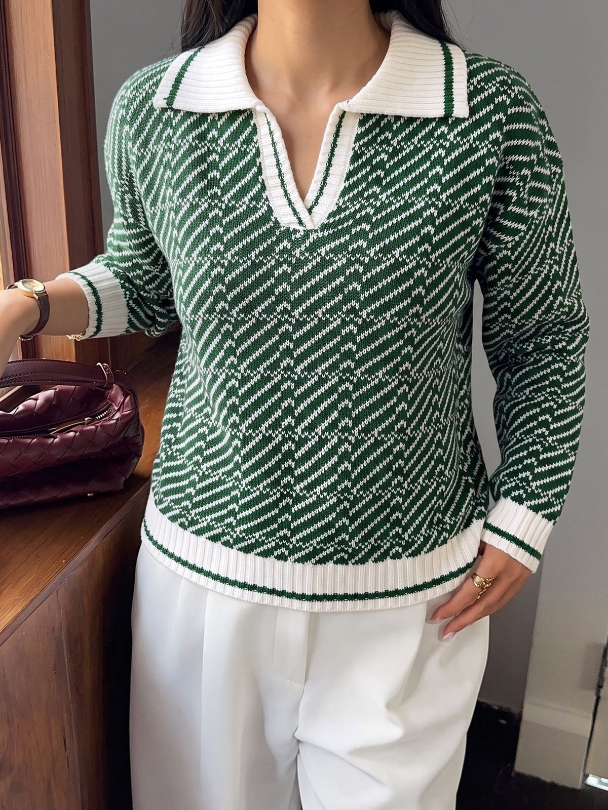 A woman wearing a green and white patterned knit polo sweater with white pants, standing indoors near a wooden cabinet and a burgundy handbag.