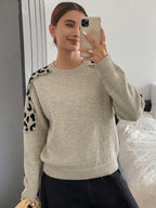 A woman takes a mirror selfie in a bedroom while wearing a light beige crewneck sweater with black leopard print patches on the shoulders, paired with a black skirt.
