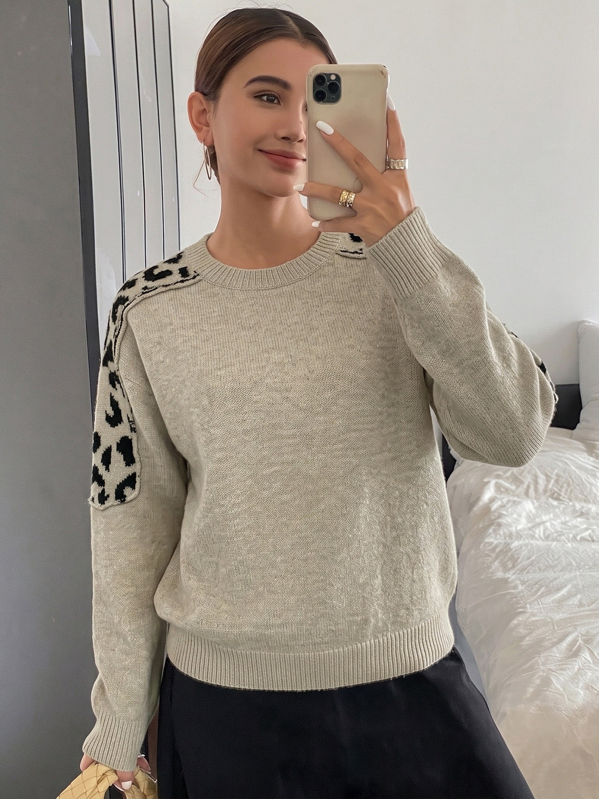 A woman takes a mirror selfie in a bedroom while wearing a light beige crewneck sweater with black leopard print patches on the shoulders, paired with a black skirt.