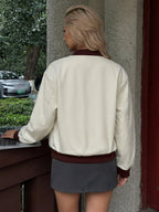 A woman viewed from behind wearing a cream-colored leather bomber jacket with dark brown ribbed trim, paired with a gray mini skirt, standing next to a concrete pillar.