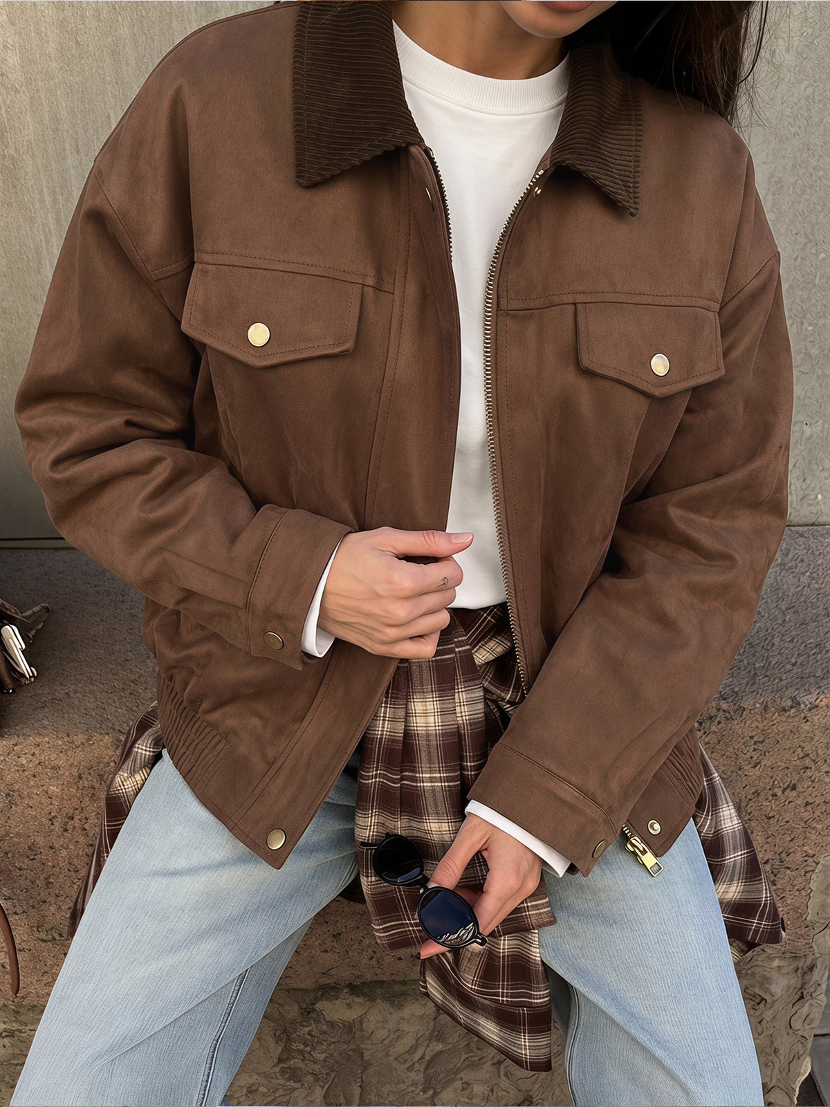 A person wearing a brown suede-like jacket with a corduroy collar, layered over a white top and light blue jeans, holding sunglasses in one hand.