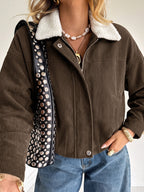 A woman wearing a brown corduroy jacket with a white sherpa collar, paired with blue jeans and gold jewelry, carrying a black studded shoulder bag.