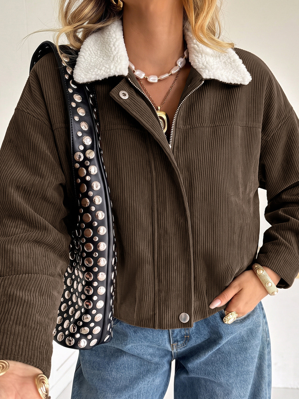 A woman wearing a brown corduroy jacket with a white sherpa collar, paired with blue jeans and gold jewelry, carrying a black studded shoulder bag.