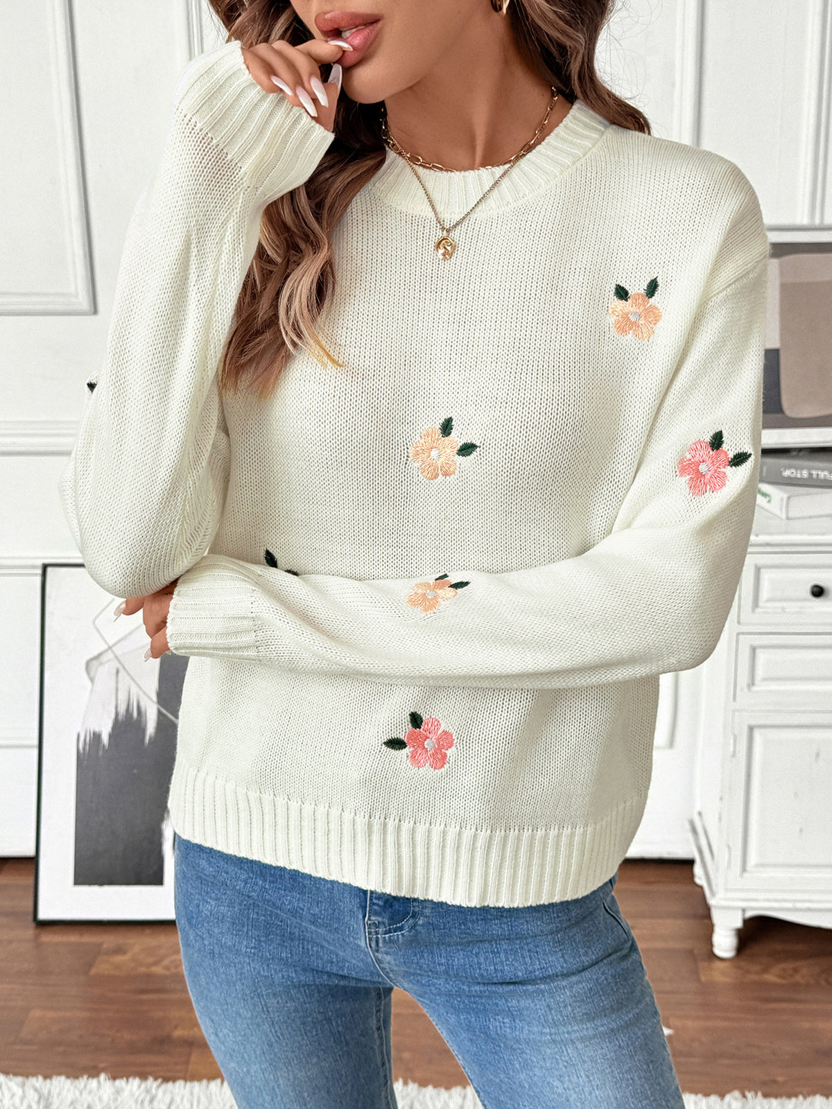 Woman wearing a white sweater with floral embroidery in a room.