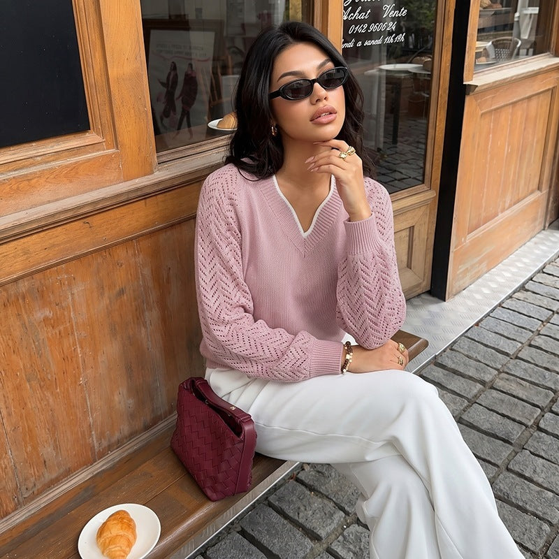 A woman sitting on a wooden bench outside a cafe, wearing a dusty pink V-neck knit sweater with white wide-leg trousers, black sunglasses, and a burgundy quilted shoulder bag.
