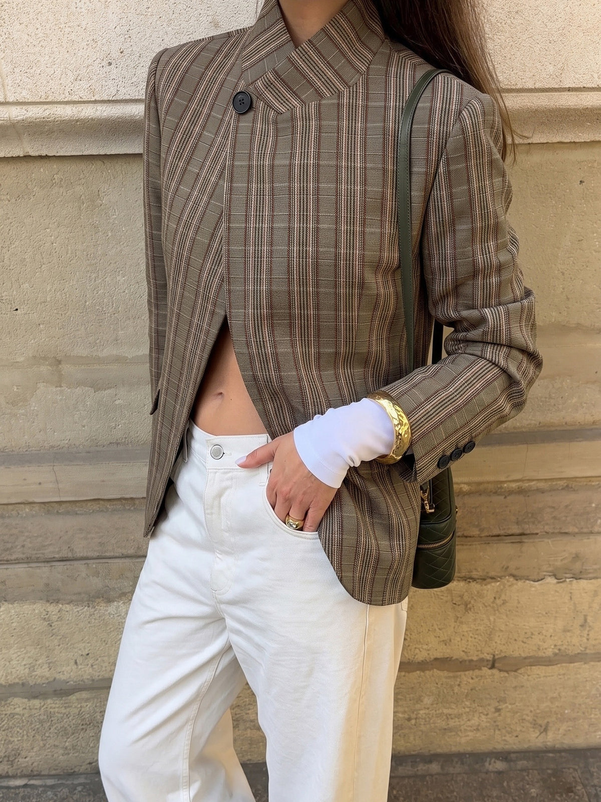 A person wearing a brown and beige plaid blazer with a unique asymmetrical button closure, paired with white trousers and a dark green shoulder bag, standing against a stone wall.