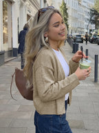 A woman with blonde hair stands on a city sidewalk wearing a beige knit cardigan over a white top and blue jeans, holding a green drink and carrying a brown shoulder bag.