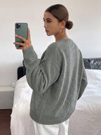 A woman takes a mirror selfie in a bedroom while wearing an oversized light gray knit sweater and white pants.
