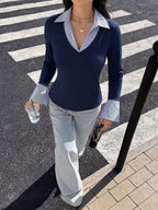 A woman walking on a crosswalk wearing a navy blue long-sleeve V-neck sweater layered over a light blue collared shirt, paired with light-wash wide-leg jeans.