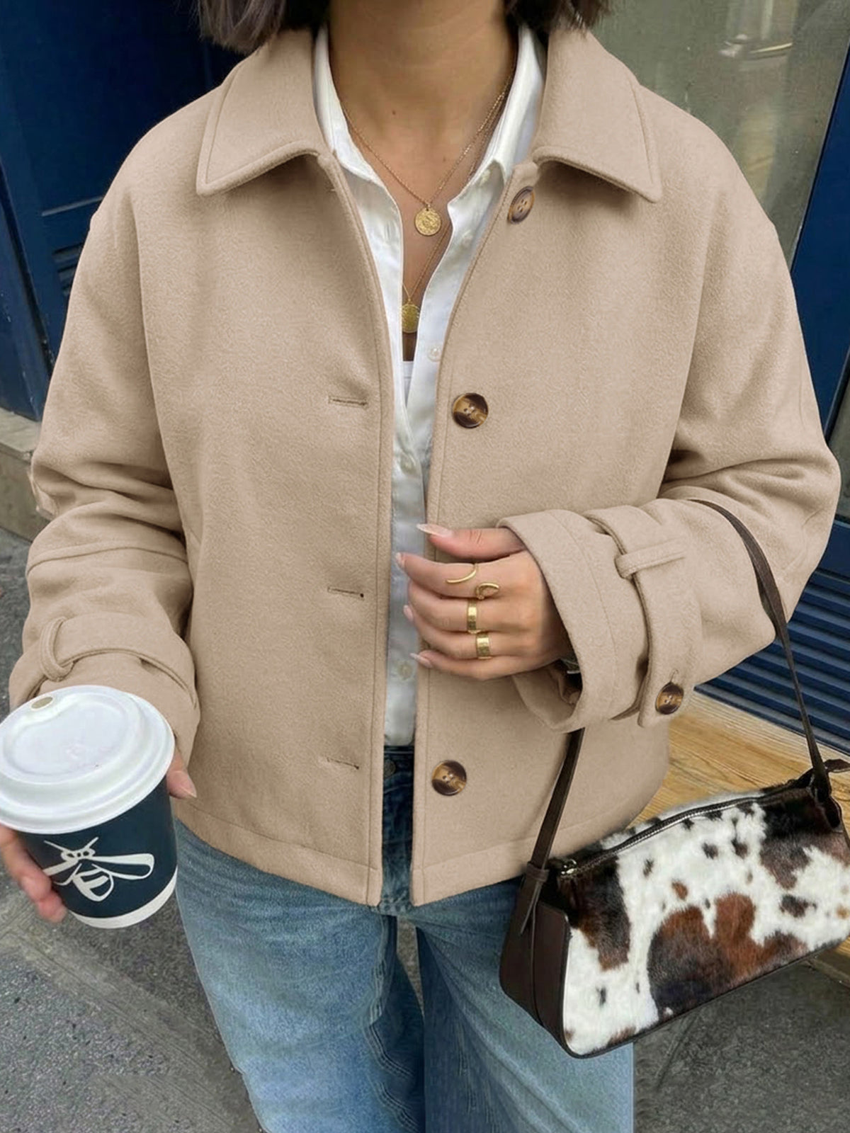 A person wearing a beige button-front coat over a white collared shirt and blue jeans, holding a coffee cup and carrying a brown-and-white cow-print shoulder bag.