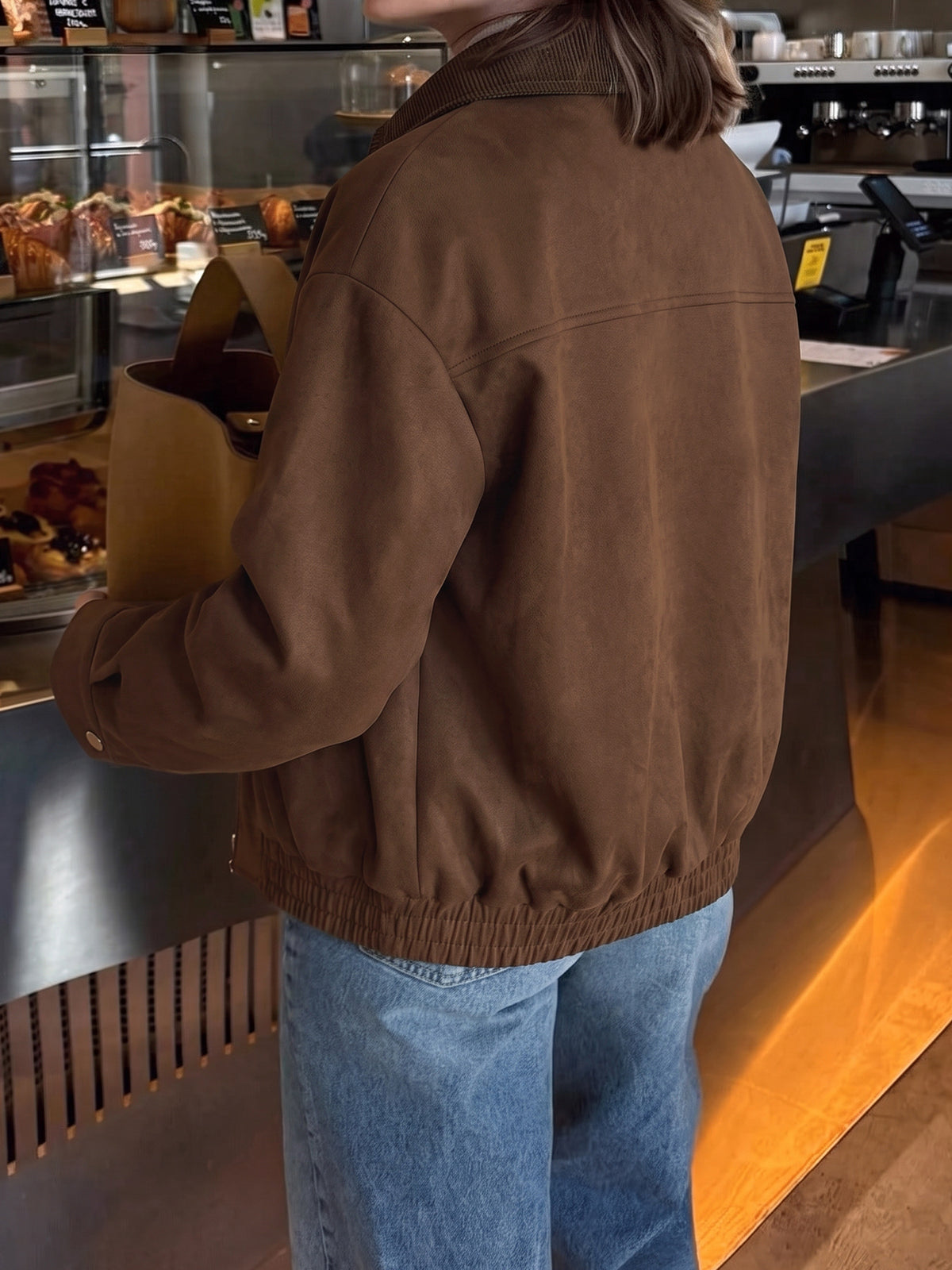 A person viewed from behind wearing a brown suede-like jacket and blue jeans, standing at a cafe counter with a paper bag in hand.