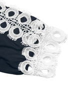 A close-up of a black garment featuring a wide white eyelet lace trim with circular cutout patterns and fringed edges, shown against a plain white background.