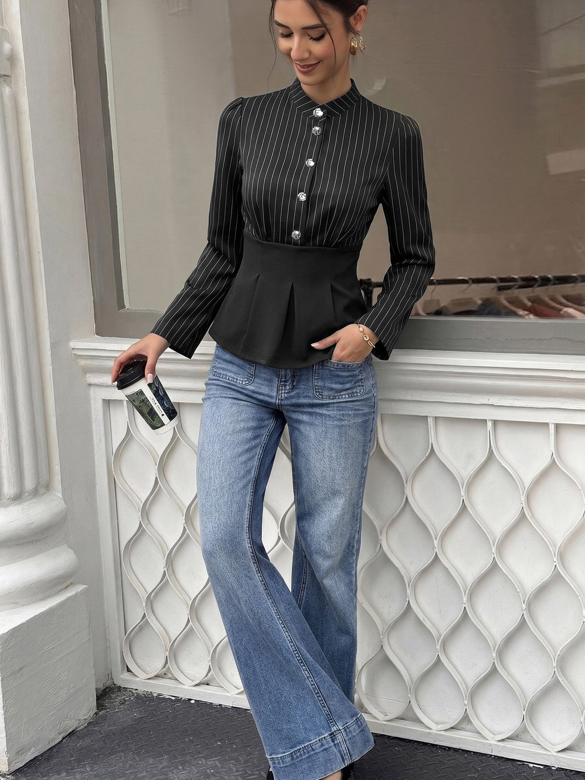 A woman models a black long-sleeve pinstriped blouse with a high collar and buttoned waist, paired with light-wash blue flare jeans, standing in front of a white decorative storefront window.