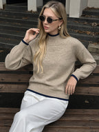 A woman wearing a beige knit mock-neck sweater with dark navy ribbed trim, paired with white trousers, sitting on a wooden bench outdoors.