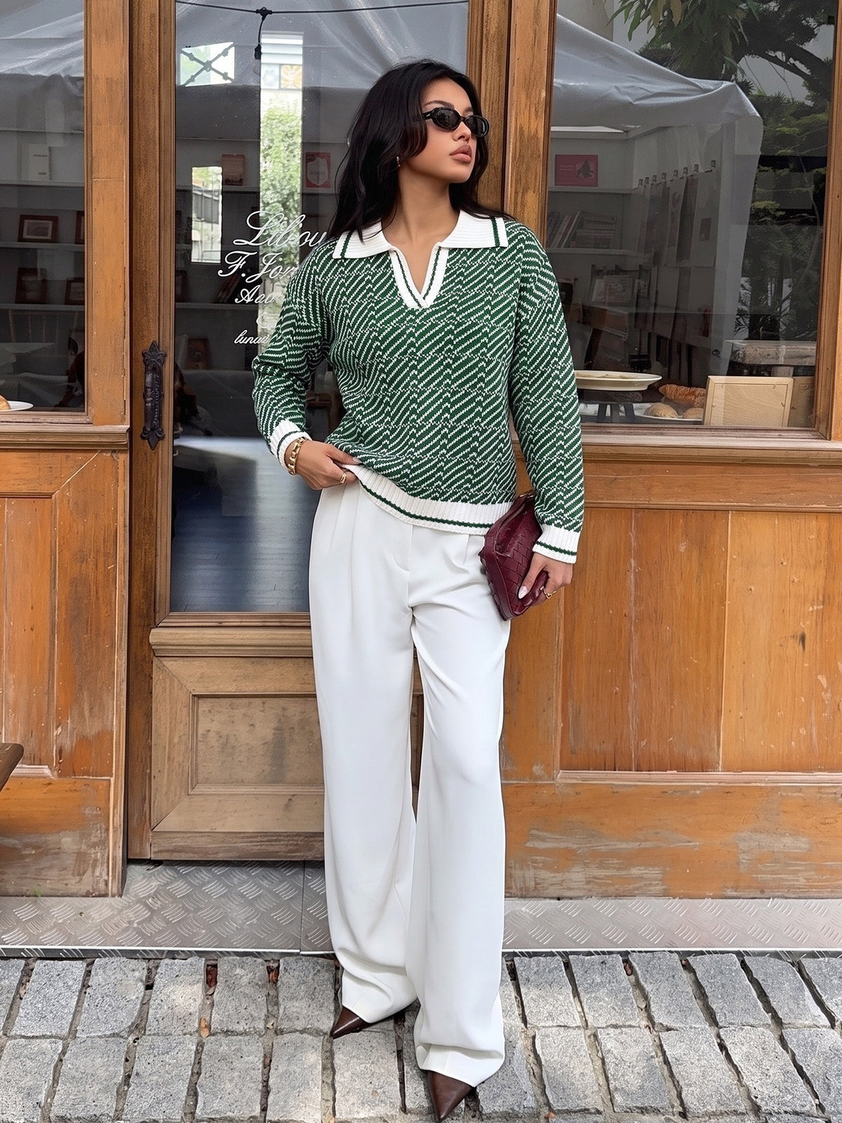 A woman stands in front of a wooden storefront wearing a green and white chevron-patterned knit sweater with a white collar and cuffs, paired with white wide-leg pants and brown pointed-toe heels. She is holding a small burgundy clutch.