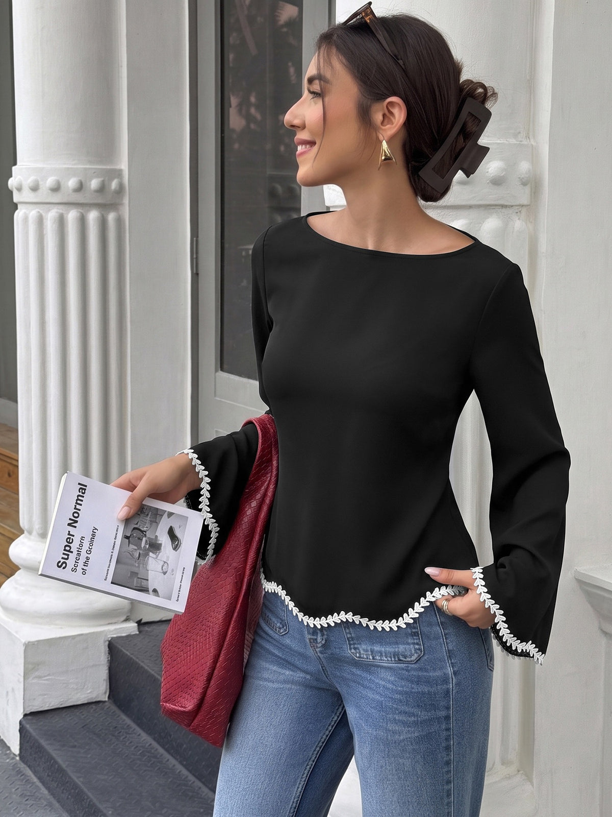 A woman wearing a black long-sleeve blouse with white pearl trim at the cuffs and hem, paired with blue jeans, holding a red textured clutch and a magazine in front of a white classical building facade.