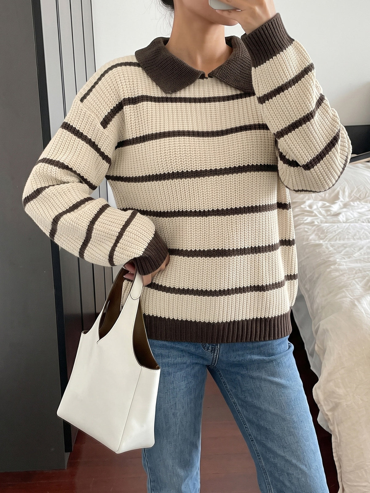 A person wearing a cream and dark brown horizontally striped knit sweater with a dark brown collar, paired with blue jeans and holding a white tote bag.