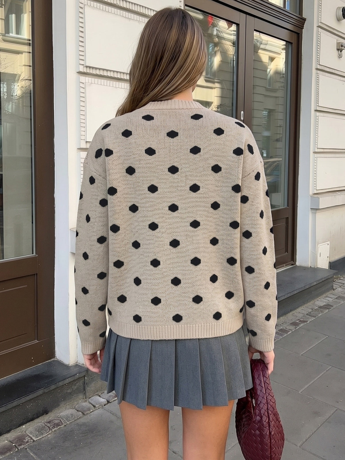 A woman viewed from behind wearing a beige knit sweater with black polka dots and a gray pleated skirt, standing on a city sidewalk in front of a building.