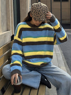 A person sitting on a wooden bench outdoors, wearing a fuzzy striped sweater with blue, yellow, and brown horizontal bands, light blue jeans, and a leopard-print cap while holding an iced coffee.