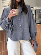 A woman wearing a blue and white gingham button-up blouse with dramatic tie details on the sleeves, paired with cream-colored pants, holding a ceramic mug in a cafe setting.
