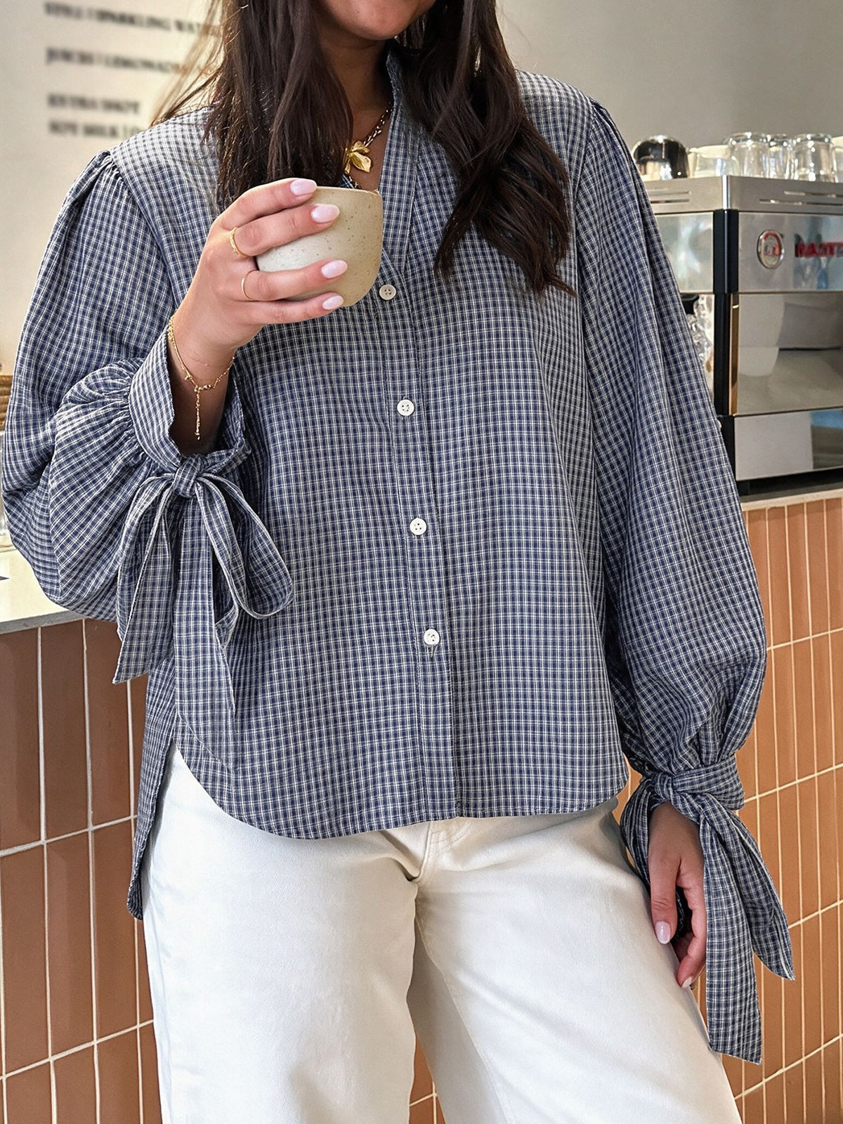 A woman wearing a blue and white gingham button-up blouse with dramatic tie details on the sleeves, paired with cream-colored pants, holding a ceramic mug in a cafe setting.