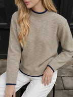 A woman wearing a beige knit sweater with dark navy blue ribbed trim at the crew neck, cuffs, and hem, paired with white pants.