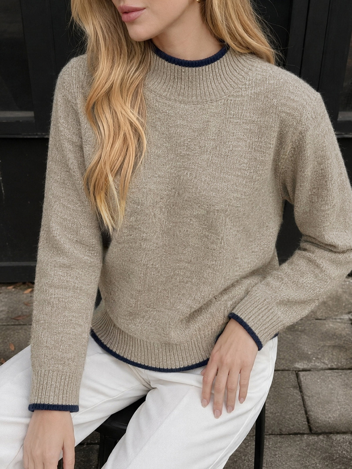 A woman wearing a beige knit sweater with dark navy blue ribbed trim at the crew neck, cuffs, and hem, paired with white pants.