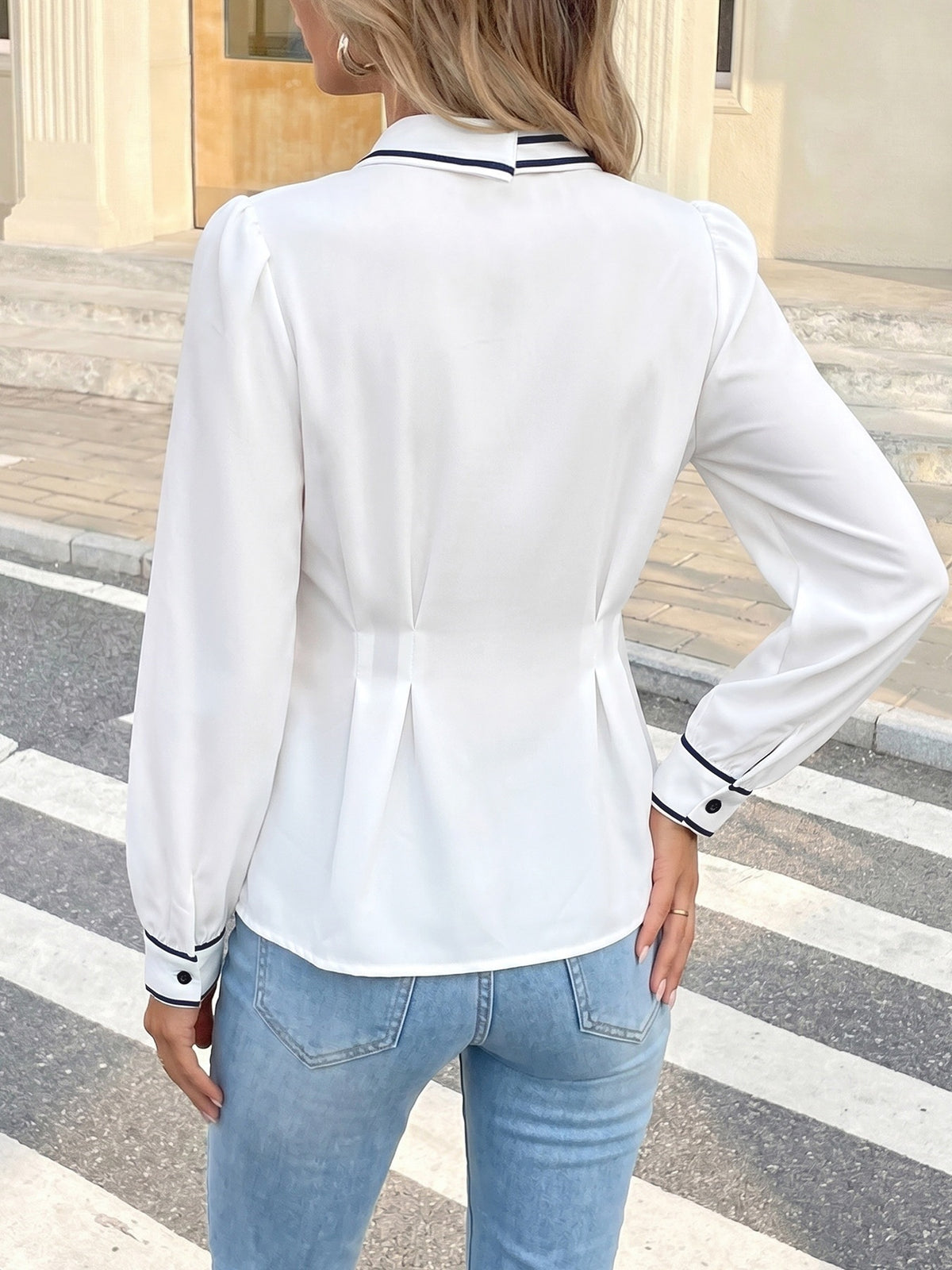A woman shown from the back wearing a white long-sleeve blouse with navy and white striped trim on the collar and cuffs, paired with light blue jeans, standing on a street crosswalk.