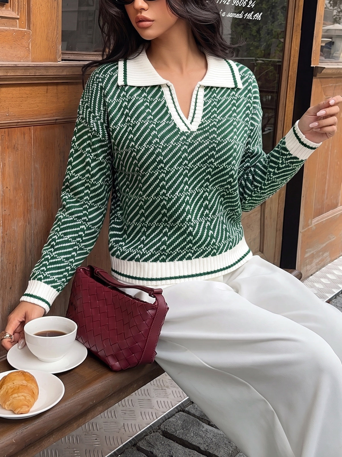A woman wearing a green and white patterned knit sweater with a white collar, seated outdoors next to a burgundy woven handbag, a cup of coffee, and a croissant.