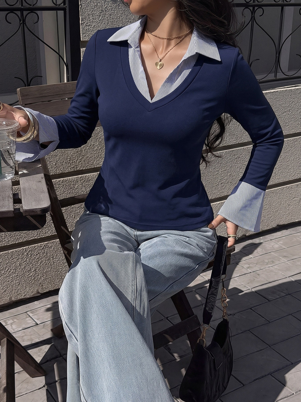 A woman seated outdoors wearing a fitted long-sleeve V-neck top with a contrasting light blue collar and cuffs over a navy body, paired with light blue jeans.