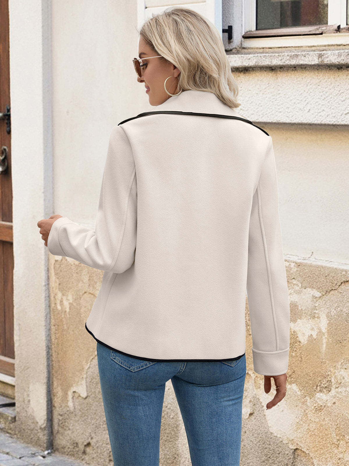 Woman wearing a beige jacket with black trim, standing against a light-colored wall.