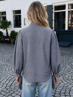 A woman viewed from behind wearing a navy and beige gingham long-sleeve blouse with balloon sleeves and side tie details, paired with light blue jeans, standing on a cobblestone courtyard in front of a white building.