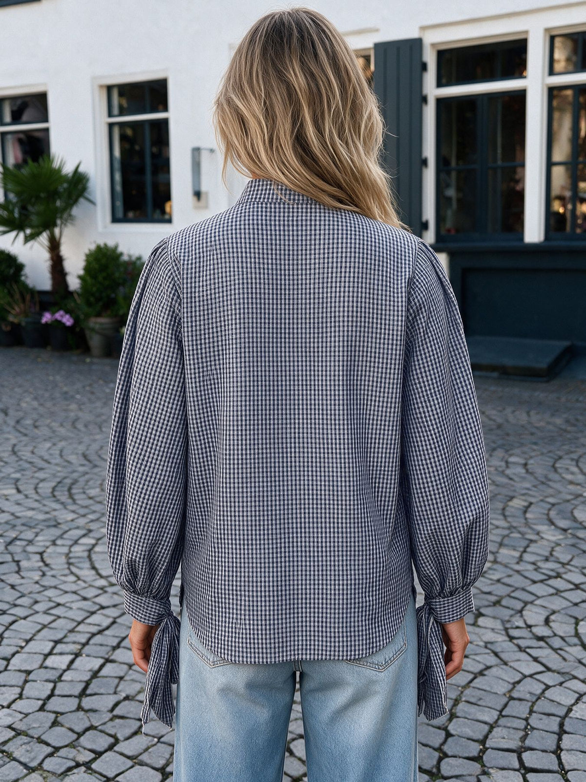 A woman viewed from behind wearing a navy and beige gingham long-sleeve blouse with balloon sleeves and side tie details, paired with light blue jeans, standing on a cobblestone courtyard in front of a white building.