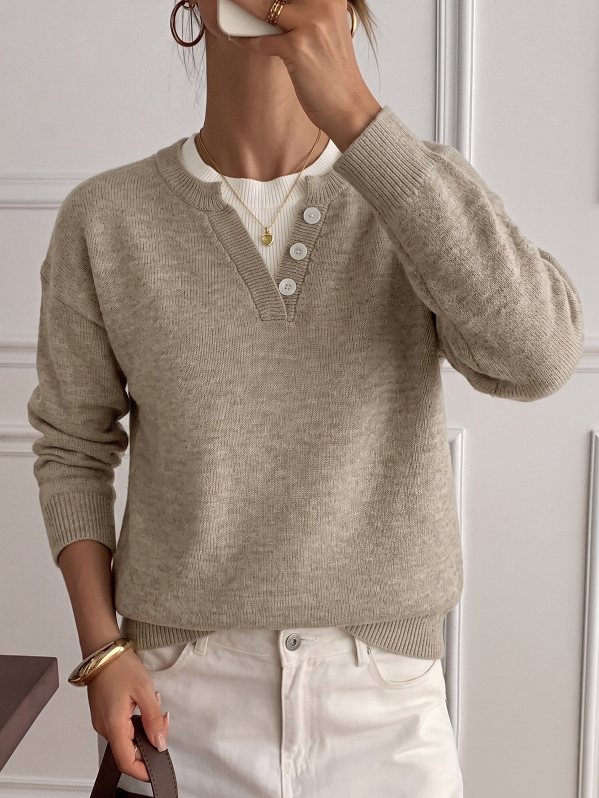 A woman wearing a light beige knit sweater with a split V-neck and three white buttons, layered over a white ribbed top and paired with white pants.
