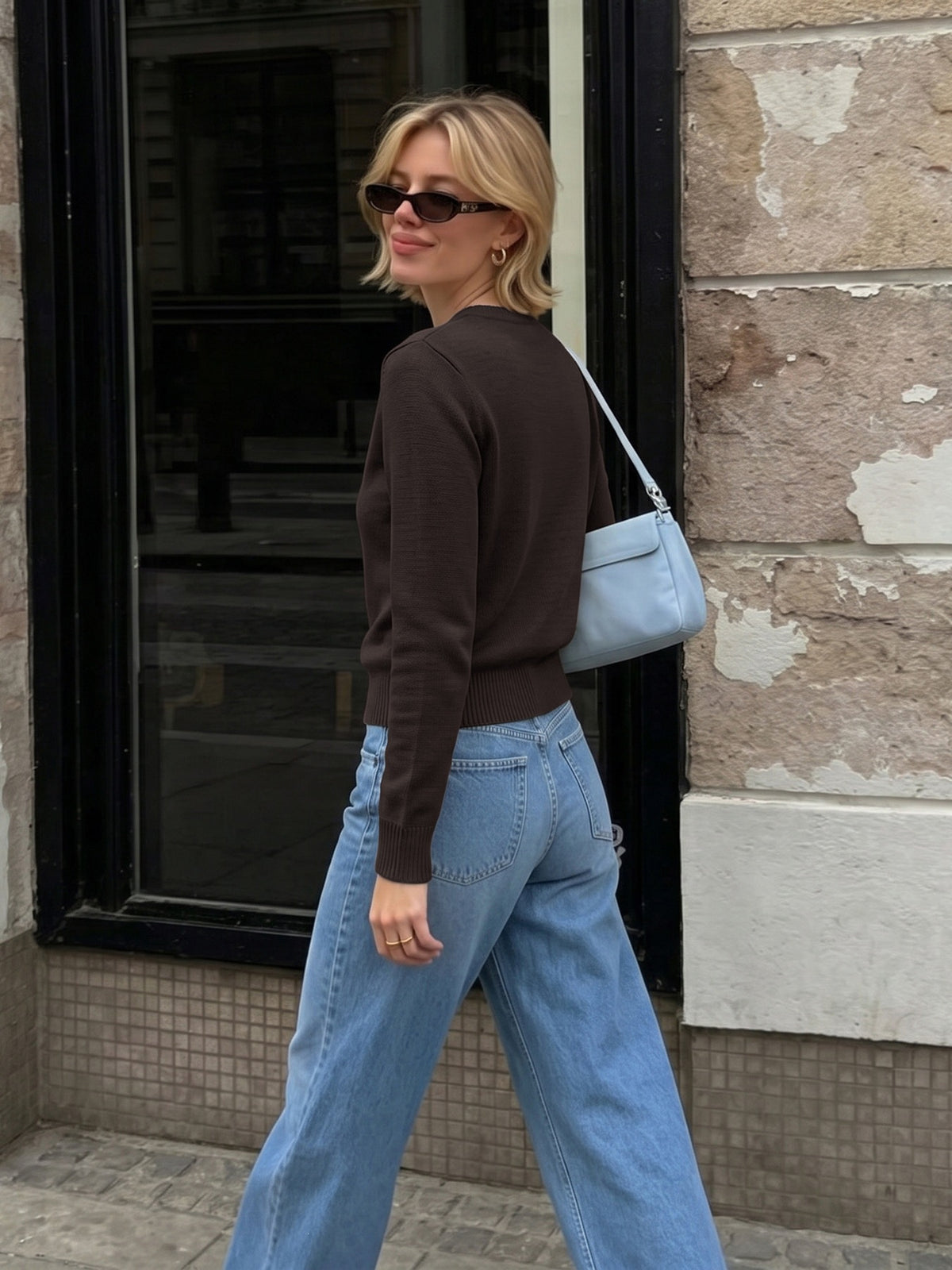 A woman wearing a dark brown long-sleeve knit top, light blue wide-leg jeans, and a light blue shoulder bag walks past a stone building.