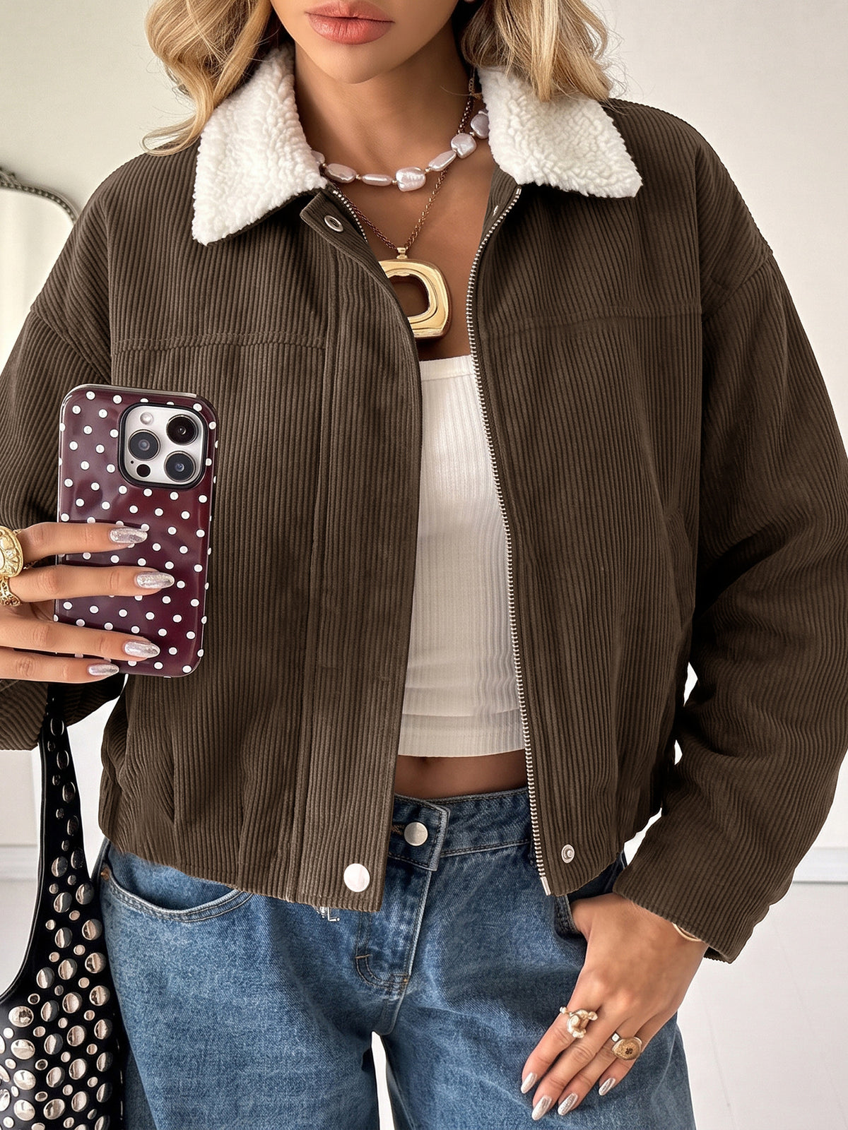 A woman wearing a brown corduroy jacket with a white sherpa collar, paired with a white ribbed tank top and blue jeans, holding a polka-dot phone case.