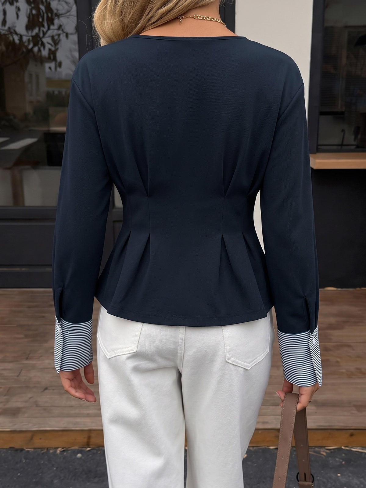 A woman viewed from the back wearing a navy long-sleeve peplum top with striped buttoned cuffs and white pants.