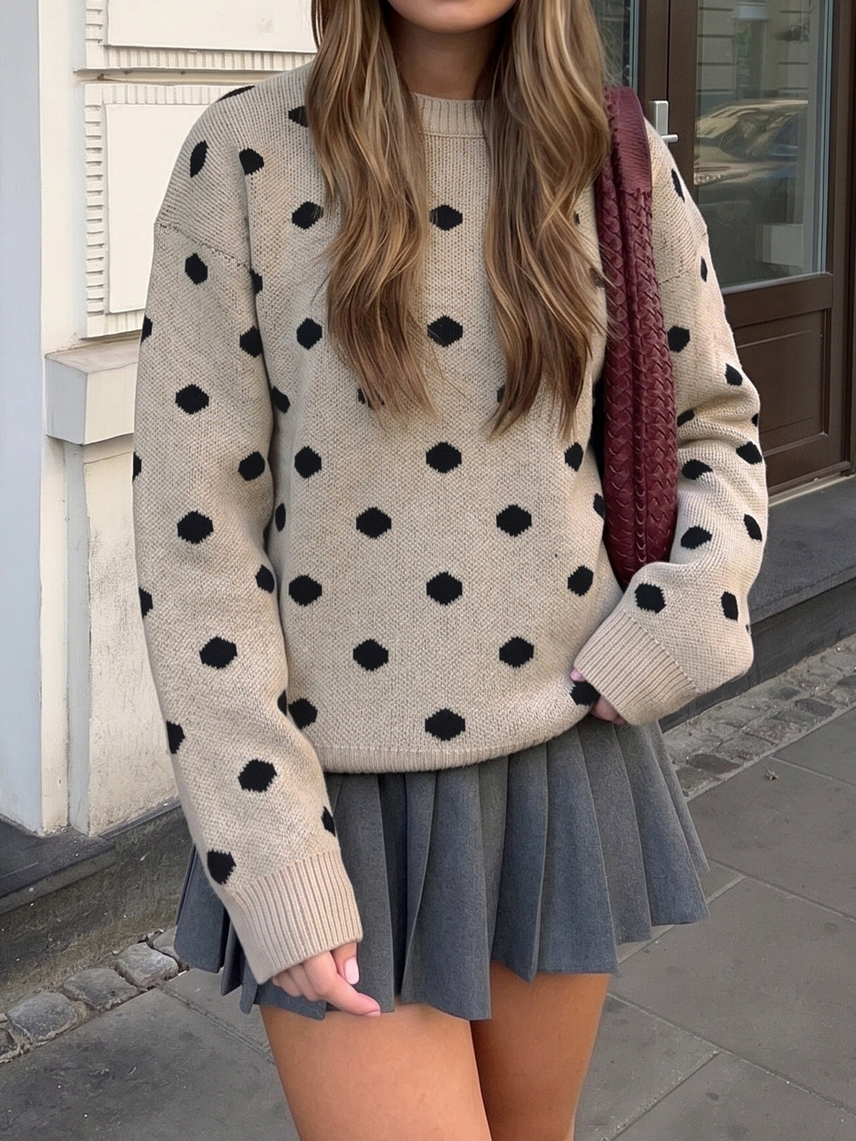 A woman wearing a beige knit sweater with black polka dots and a gray pleated mini skirt, carrying a burgundy knit shoulder bag, standing outdoors near a building entrance.