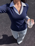 A woman wearing a navy blue long-sleeve V-neck top with a light blue and white striped collar, paired with light-wash jeans, holding a clear plastic cup with a green straw on an asphalt surface.