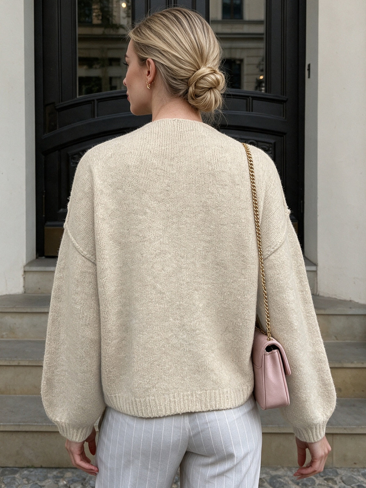 A woman viewed from behind wearing a cream-colored, textured knit sweater with balloon sleeves, paired with light gray pinstripe trousers and a small pink shoulder bag.