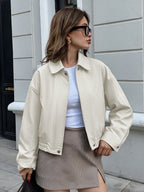 A woman wearing an oversized cream-colored jacket over a white top and a brown-and-white houndstooth mini skirt, standing outdoors against a light gray building wall.
