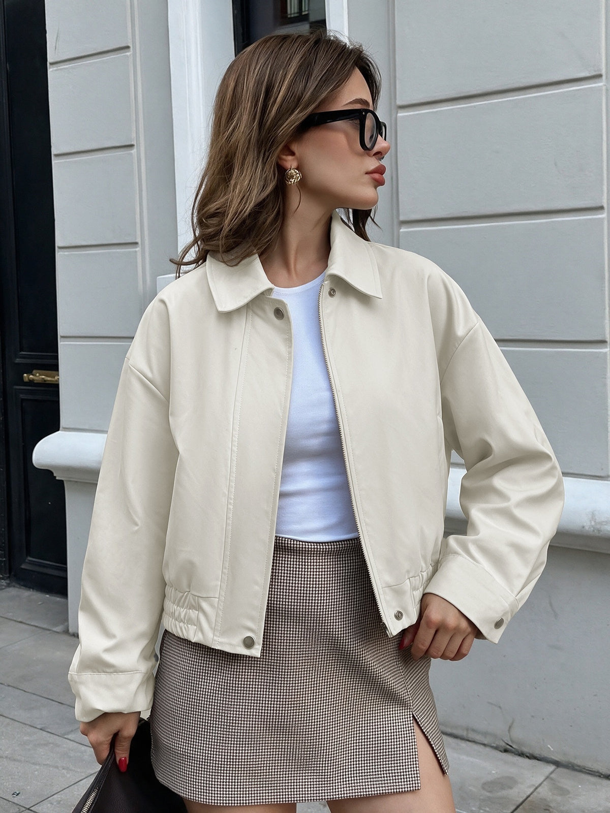 A woman wearing an oversized cream-colored jacket over a white top and a brown-and-white houndstooth mini skirt, standing outdoors against a light gray building wall.